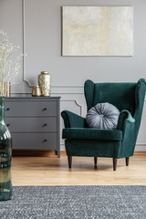 Emerald green armchair with pillow next to grey wooden commode in dark living room interior
