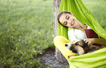 Smiling young woman resting in green hammock with cute dog Welsh Corgi in a park outdoors....
