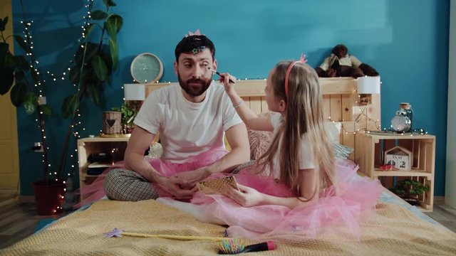 Fairy dressed cheerful father and daughter playing together at home. Little girl doing makeup to her daddy preparing for birthday party on bed.