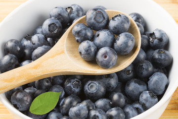 Close-up of wooden spoon with fresh ripe blueberries, against white bowl with fruits