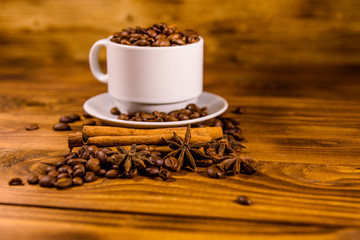White cup filled with coffee beans, star anise and cinnamon sticks on wooden table