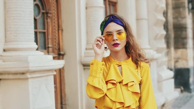 Street fashion portrait of young beautiful elegant lady wearing trendy yellow color sunglasses, headband, stylish blouse with jabot, posing in old european city