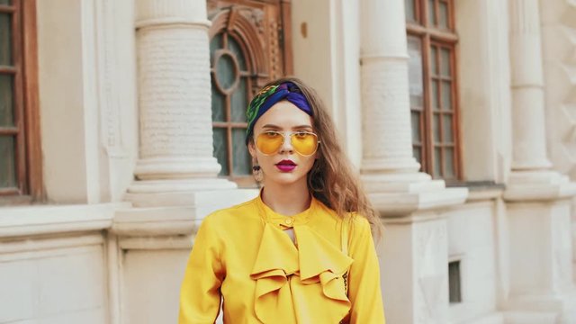 Street fashion portrait of young beautiful elegant woman wearing trendy yellow color aviator sunglasses, headband, stylish blouse with jabot, posing in old european city