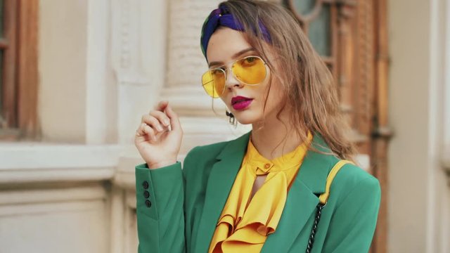 Street fashion portrait of young beautiful elegant lady wearing trendy yellow color sunglasses, headband, stylish blouse, green blazer, posing in old european city