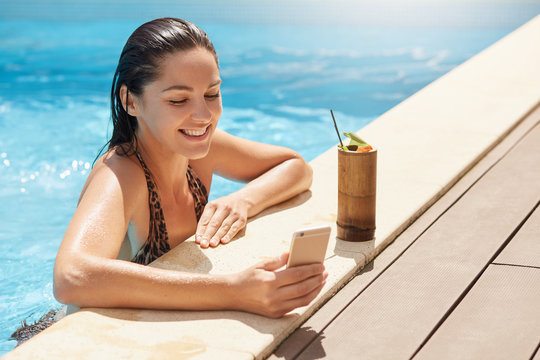 Image of cheerful delighted model having pleasant facial expression, taking photo in swimming pool, smiling sincerely, being in high spirits, having break with cocktail, talking through videochat.