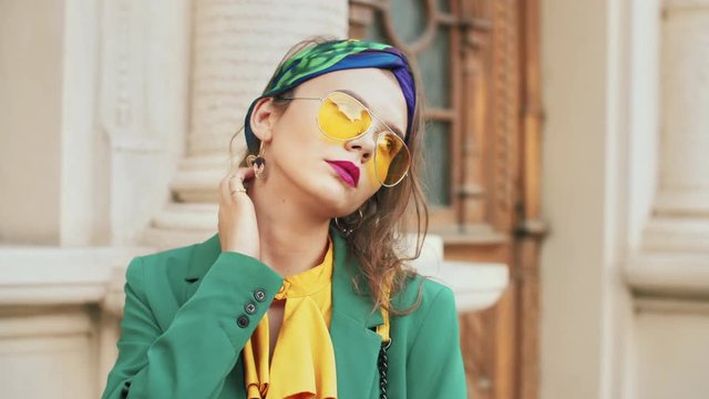 Street fashion portrait of young beautiful elegant woman wearing trendy yellow color sunglasses, headband, stylish blouse, green blazer, posing in old european city