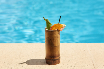 Close up picture of wooden container with cocktail situated near swimming pool on ground. There are black straw, green leaf, slices of orange and grapefruit inside cup. Summer drinks concept.