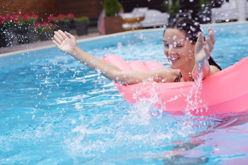Joyful funny model splashing water in swimming pool, swimming with mattress, raising her hands, having fun in water, enjoying her rest at spa resort, chilling out alone. Vacation time concept.