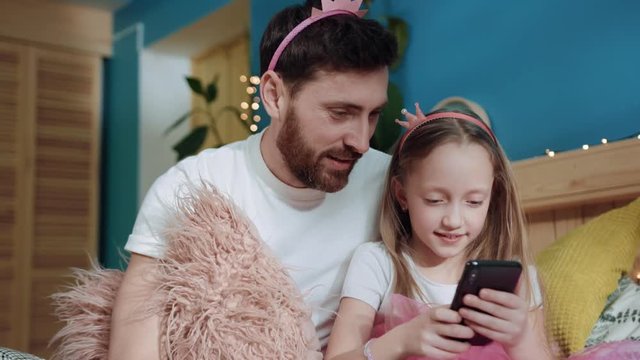 Closeup portrait excited cute girl leaning on her father dressed like fairies watching cartoons together om smartphone sitting on bed at home. Party celebration birthday.