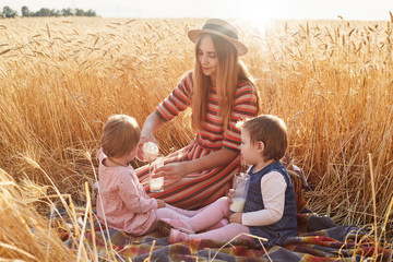 Caring helpful mother sitting on blanket at wheat field with her lottle daughters, filling glasses with milk from bottle, wearing dress and hat, childen behaving in proper way. Motherhood concept.