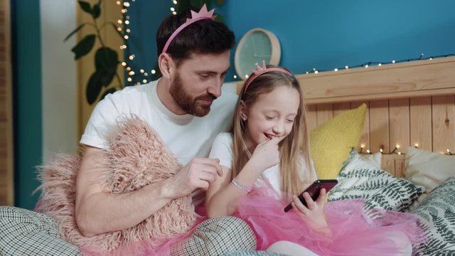 Beautiful family portrait of cheerful daddy and his little girl using a smartphone while sitting on bed. Father and daughter dressed like magic fairies playing together at home. Family party