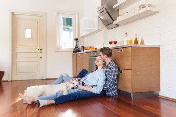 Fototapeta premium Happy young married couple resting sitting on the floor near the kitchen set hugging their beloved dog. Holiday concept during the weekend