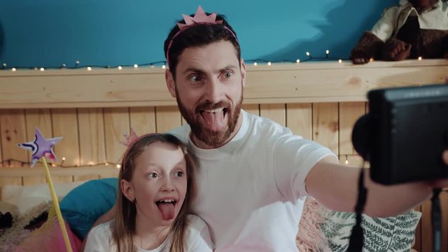 Closeup portrait funny daddy and his daughter little princess showing silly grimace with tongues out taking joint selfie in bedroom at home.
