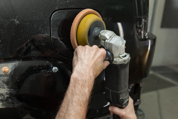 Close-up view on the hands of a male worker who holds a tool for polishing the fender of a car while working in a vehicle detailing workshop. Auto service industry.