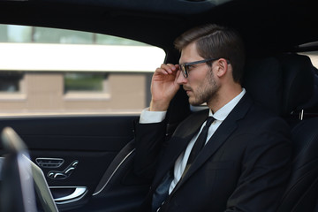 Thoughtful confident businessman keeping hand on glasses while sitting in the luxe car.