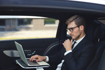 Thoughtful young businessman keeping hand on chin while sitting in the lux car and using his laptop.