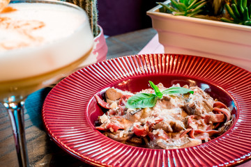Red fettuccine pasta with fried mushrooms in a creamy sauce in a red plate, and an yellow cocktail with foam in a beautiful glass on a table in a restaurant. Close-up. Space