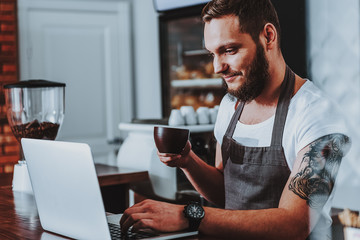 Relaxed barista drinking coffee and using his laptop