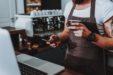 Barista standing with coffee and using modern smartphone