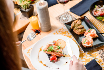 Young woman eating roasted salmon fillet and risotto with cuttlefish ink on a white plate with a signature sauce and Cherry tomatoes in a restaurant. Brunch friends in the restaurant. From the back