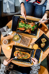 Two girls have breakfast in the restaurant. Spring rolls and grilled meat with pumpkin puree on black plates on a light wooden table in a restaurant. Flat lay. top view