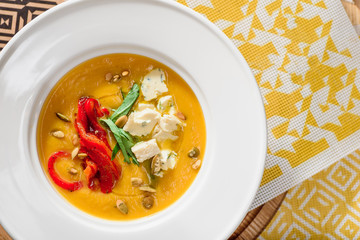 pumpkin cream soup with cheese Dor Blue, bell pepper, pumpkin seeds in a white plate on yellow set on a wooden table in a restaurant. Pumpkin soup with toast on a table in a restaurant. close-up