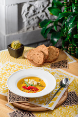 pumpkin cream soup with cheese Dor Blue, bell pepper, pumpkin seeds in a white plate on yellow set on a wooden table in a restaurant. Pumpkin soup with toast on a table in a restaurant. close-up