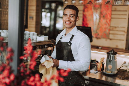 Good-looking barman wiping white cup with cleaning cloth