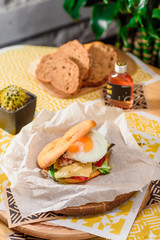 Burger with fried egg, chicken, cheese, tomatoes and other vegetables on a craft paper on yellow background in a restaurant. Toasted black bread on the background. Series of photos for the menu.