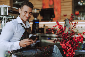 Handsome barman in black apron using cellphone in cafe