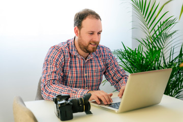 Young male Photographer with blue eyes, redhead beard and plaid shirt working from home with a laptop sitting next to a green plant. Photo camera on a table. Happy worker on his own.