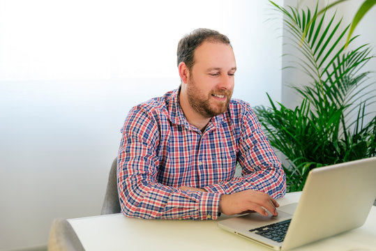 Real man with blue eyes and redhead Beard dressed in plaid shirt Working with a computer from home next to a green plant and window in the background. Self-employed. Technological works IT