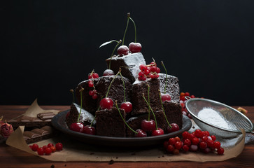Homemade brownie (cake) on parchment paper. Near cherry, icing sugar and red currants. Black background