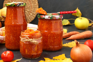 Homemade adzhika with tomatoes, apples and carrots in jars on a dark background, harvesting for the winter