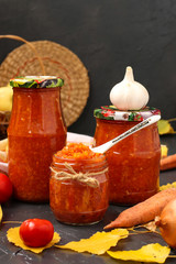 Homemade adzhika with tomatoes, apples and carrots in jars on a dark background, harvesting for the winter, vertical