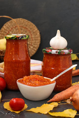 Homemade adzhika with tomatoes, apples and carrots in jars on a dark background, harvesting for the winter, vertical