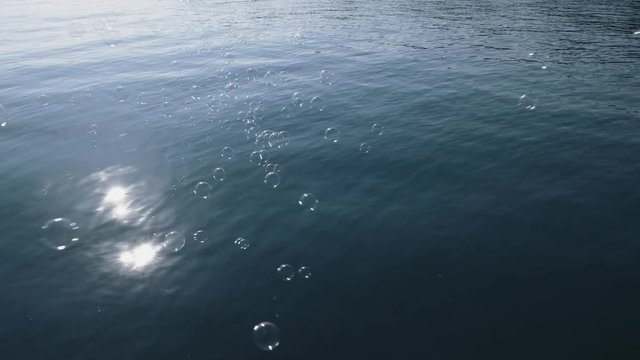 Slow motion on the view of the texture of the blue sea with small waves and flying soap bubbles on the surface of the water. Reflection of the sun disk in water and rays of light. Ready footage