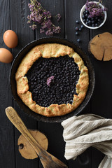 Homemade comfort food rustic style. Crusty blueberry galette in cast iron pan with ingredients and marjoram on dark background