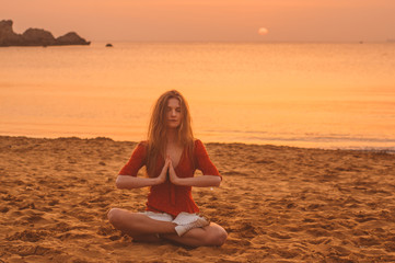 Fototapeta na wymiar Young blonde girl in red dress sitting in Lotus position in the evening on the beach