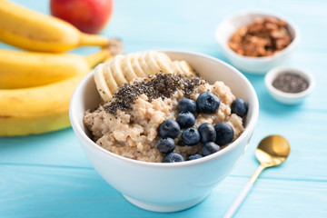 Oatmeal porridge with banana, blueberries and chia seeds in white bowl on blue background. Healthy breakfast food
