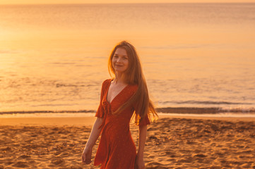 Fototapeta na wymiar Young blonde girl in red dress walking in the evening on the beach