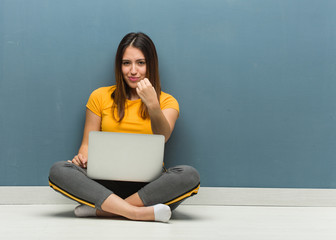 Young woman sitting on the floor with a laptop showing fist to front, angry expression