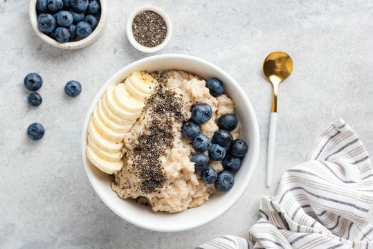 Breakfast oatmeal porridge with banana, blueberries, chia seeds on grey concrete background. Table top view of healthy food for weight loss, clean eating, vegan and vegetarian diet