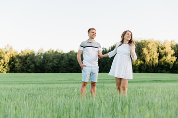 Naklejka na ściany i meble Young pretty couple in love standing in the field. Handsome cheerful blonde girl in white dress hugging her boyfriend. Man and woman having fun outdoors