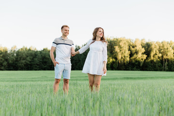 Young pretty couple in love standing in the field. Handsome cheerful blonde girl in white dress hugging her boyfriend. Man and woman having fun outdoors