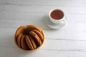 a cup of tea and a bowl of oatmeal cookies on white wooden background