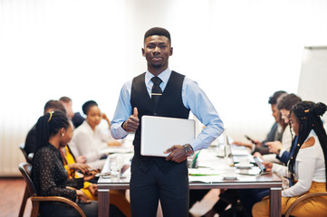Face of handsome african business man, holding laptop and show thumb up on the background of business peoples multiracial team meeting, sitting in office table.