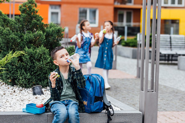 Fototapeta premium Boy eating outdoors the school from plastick lunch boxe. Healthy school breakfast for child. Food for lunch, lunchboxes with sandwiches, fruits, vegetables, and water.