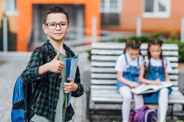 Funny little boy in glasses. Child from elementary school with book and bag. Okay sign.