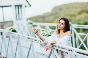 Smiling young female or bride posing with cup of some drink, wearing at white robe. Morning concept.